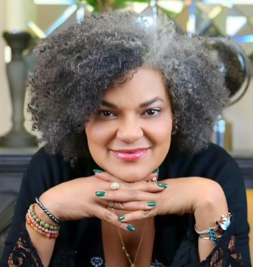 Woman with curly gray hair smiling, hands clasped in front, wearing bohemian-style clothing and jewelry, representing empowerment and authenticity in intuitive coaching.