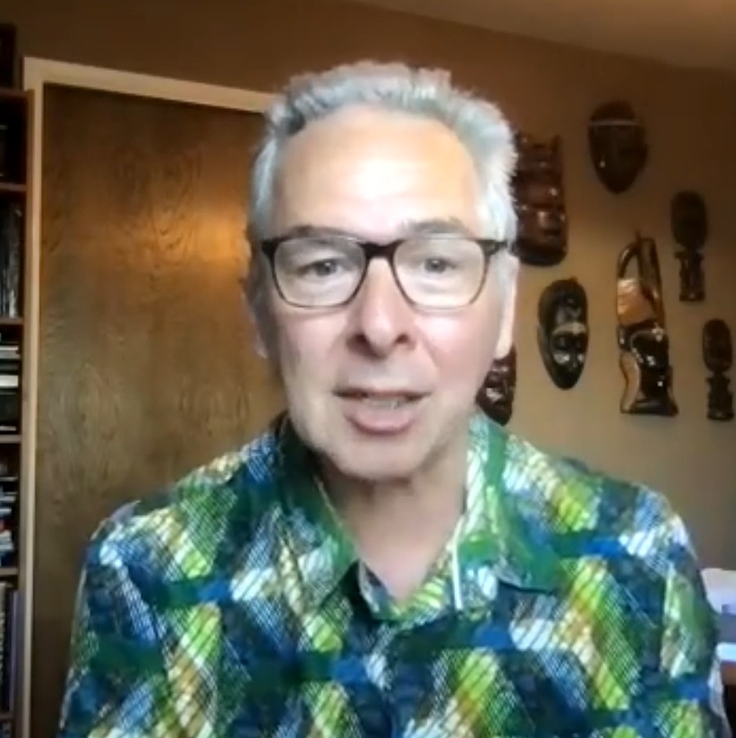 Jim Larkin, smiling and speaking, wearing a colorful plaid shirt, with wooden masks displayed on the wall behind him, representing the supportive community of Elevate coaching sessions.